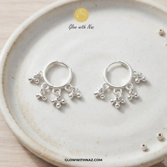 12 mm Anti-Tarnish Silver 3 Drop Separate Hoops Earrings for Women - 12 mm Anti-Tarnish Silver 3 Drop Separate Hoops Earrings for WomenJewelryGlow With NazGlow With NazA pair of silver huggie hoop earrings with dangling floral and bead charms, displayed on a round beige ceramic plate. The brand logo 'Glow with Naz' is visible at the top, and the website 'GLOWWITHNAZ. Silver 3 Drop Separate Hoops, Glow With Naz earrings, stainless steel silver earrings, viral trending earrings, drop huggie hoops,