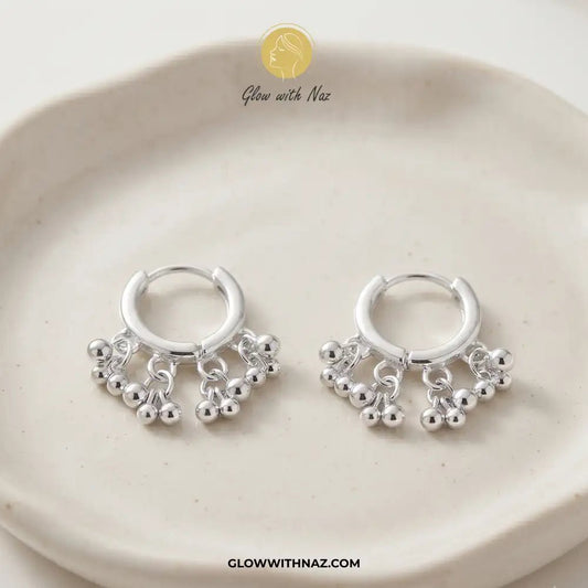 12 mm Silver 3 Attached Silver Drop Hoops for Women - 12 mm Silver 3 Attached Silver Drop Hoops for WomenJewelryGlow With NazGlow With NazE046A pair of silver huggie hoop earrings with dangling beaded fringe, displayed on a light beige ceramic dish. A gold logo for 'Glow with Naz' is visible at the top, and the website 'GLOWWITHNAZ. Silver 3 Drop Separate Hoops, Glow With Naz earrings, stainless steel silver earrings, viral trending earrings, drop huggie hoops, silver dangle earrings, eye-catchi