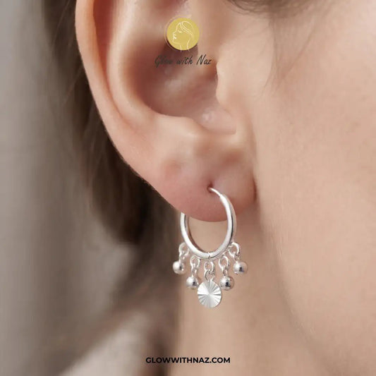 12mm Anti-Tarnish Silver 5 Drop Trending Hoops Earrings - 12mm Anti-Tarnish Silver 5 Drop Trending Hoops EarringsJewelryGlow With NazGlow With NazE045A close-up of a person's ear wearing a silver hoop earring with dangling charm beads and a central sunburst disc. The image features a watermark logo for Glow with Naz. Silver 3 Drop Separate Hoops, Glow With Naz earrings, stainless steel silver earrings, viral trending earrings, drop huggie hoops, silver dangle earrings, eye-catching drop hoops, w