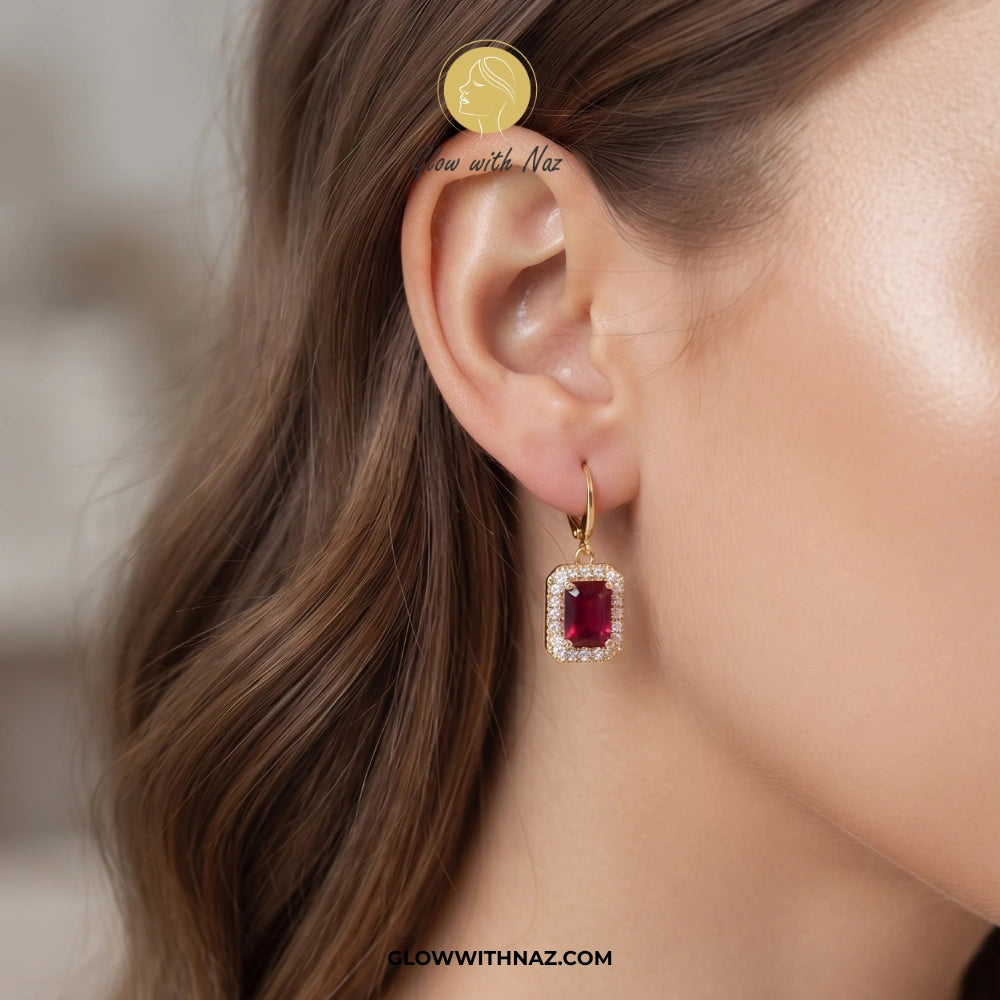 A close-up of a woman's ear wearing a gold hoop earring with a rectangular red gemstone surrounded by small clear stones. A coin is placed near the ear for scale, and the text 'Glow with Naz' appears on the image. studs earrings, small earrings, girls earrings, simple earrings, earrings for girls, black earrings, earrings, ear rings, earrings tops, stud earrings, tops earrings, aesthetic earrings, hoop earrings, drop earrings, diamond earrings, pink earrings, green earrings, ear ring, earrings for women, fl