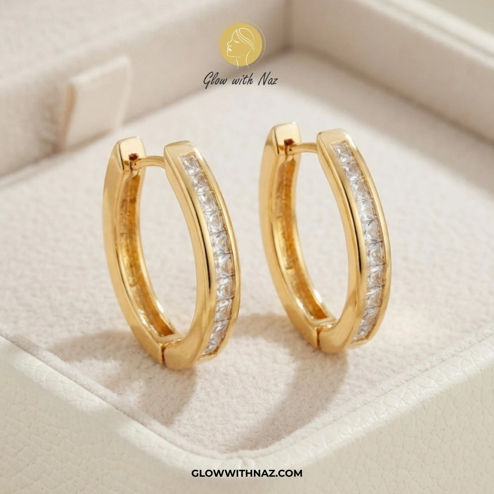 A pair of gold huggie hoop earrings with a channel-set row of clear stones on the front, displayed inside a cream-colored jewelry box. The brand logo 'Glow with Naz' is visible at the top, and the website 'GLOWWITHNAZ. studs earrings, small earrings, girls earrings, simple earrings, earrings for girls, black earrings, earrings, ear rings, earrings tops, stud earrings, tops earrings, aesthetic earrings, hoop earrings, drop earrings, diamond earrings, pink earrings, green earrings, ear ring, earrings for wome