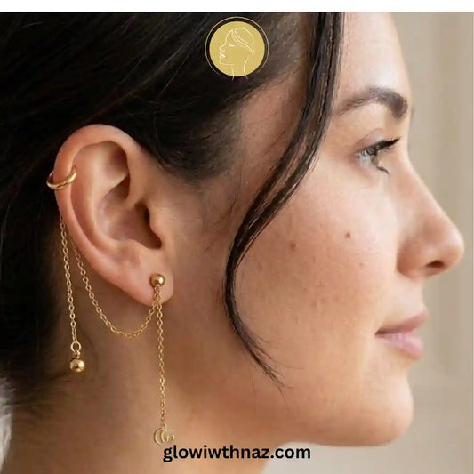 A pair of gold-toned ear piercings, including a hoop and a barbell with a dangling chain and small charm, displayed on a textured white ceramic dish. helix piercing, cartilage earring, double diamond stud, stainless steel jewelry, body jewelry, viral trending jewelry, ear stack earrings, cartilage piercing jewelry, minimalist piercing, tragus piercing, conch piercing, daith piercing, flat piercing, upper ear piercing, gold stainless steel piercing, sparkling helix stud, crystal cartilage jewelry, modern bod