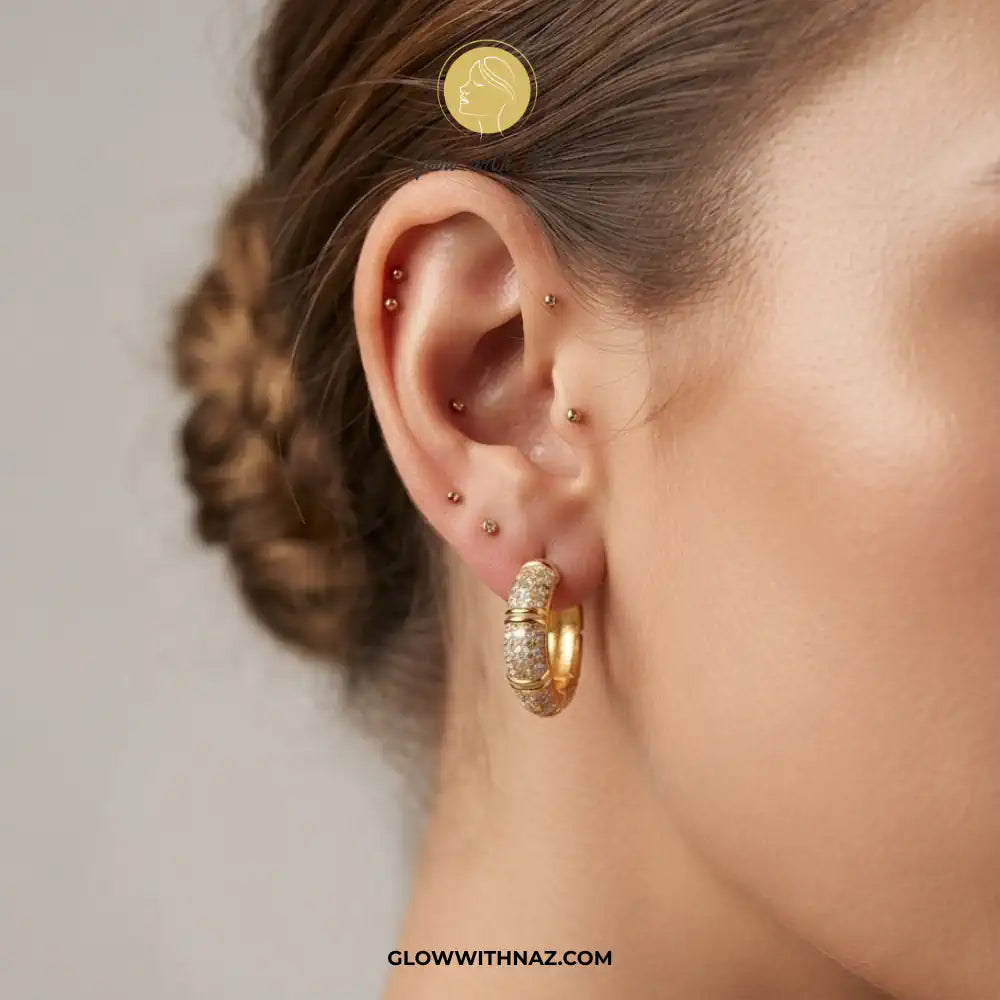 Close-up of a model's ear showcasing a textured gold huggie hoop earring, with other gold ear piercings on the lobe and cartilage. A gold coin is placed on the model's head for scale, demonstrating the earring's size.
