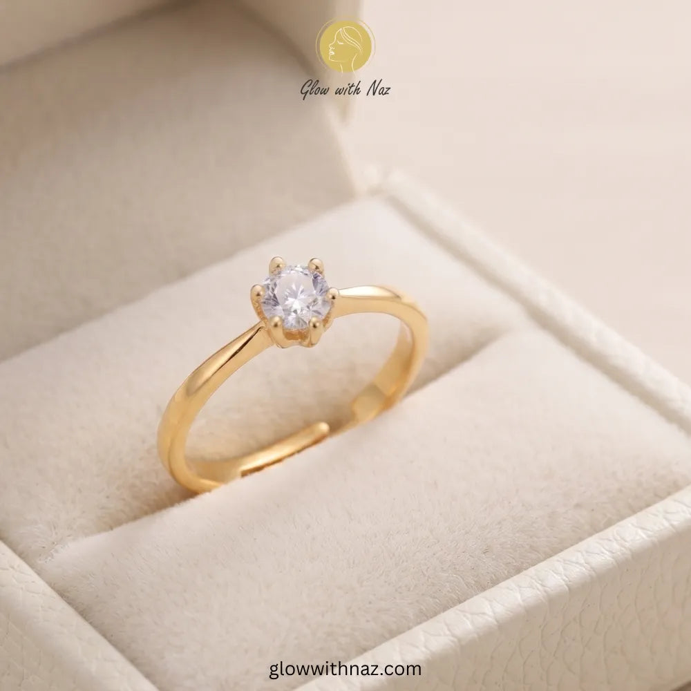 A gold ring with a single round clear gemstone in a prong setting, displayed inside a cream-colored jewelry box. The brand name 'Glow with Naz' is visible at the top, and a website URL is visible at the bottom.