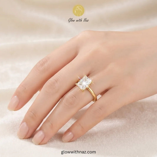 Adjustable Golden Radiant Rectangle Diamond Ring - Adjustable Golden Radiant Rectangle Diamond RingJewelryGlow With NazGlow With NazR021A gold ring with a square clear gemstone set on a person's ring finger, shown against a soft beige fabric background. The brand name 'Glow with Naz' is visible at the top. ring, girls rings, rings for girls, rings simple, simple rings, stainless steel jewelry, jewellery set, aesthetic jewelry, simple jewellery, chain, jewelry, artificial jewellery