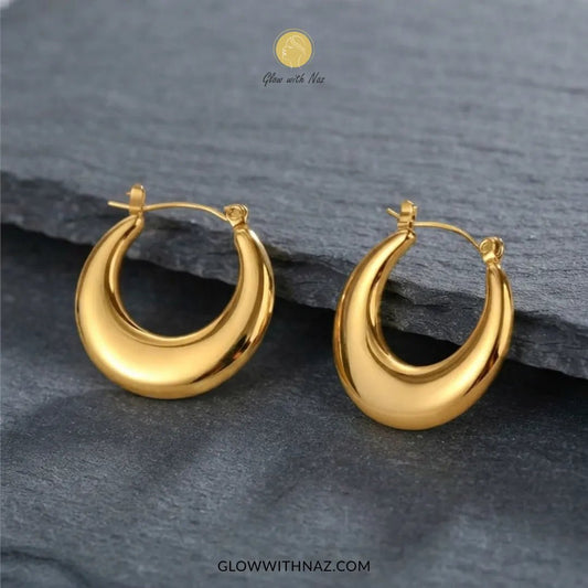 Gold Stainless Steel Plain Thick Chunky Baali Hoop Earrings - #Gold Stainless Steel Plain Thick Chunky Baali Hoop EarringsJewelryGlow With NazGlow With NazE0100A pair of polished gold crescent-shaped hoop earrings displayed on a dark slate surface. The brand logo 'Glow with Naz' is visible at the top, and the website 'GLOWWITHNAZ.#