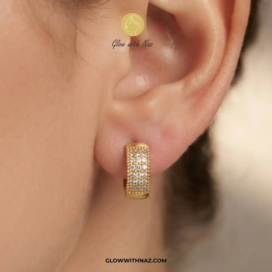 Minimal Zircon Mini Thick Hoops Earring for Girls - Minimal Zircon Mini Thick Hoops Earring for GirlsJewelryGlow With NazGlow With NazE055A close-up of a person's ear wearing a small gold huggie hoop earring set with clear pavé stones. The image includes a watermark logo for 'Glow with Naz' and the website 'GLOWWITHNAZ.