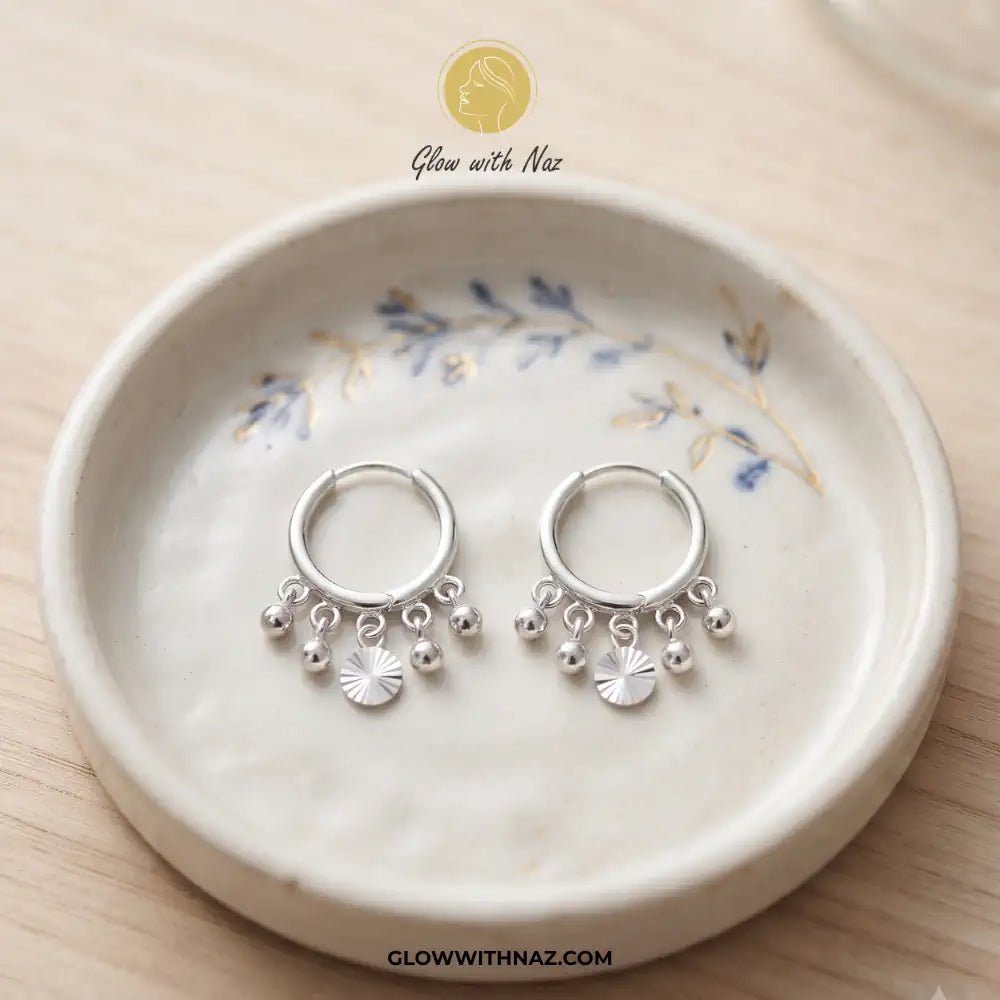 12mm Anti-Tarnish Silver 5 Drop Trending Hoops Earrings - 12mm Anti-Tarnish Silver 5 Drop Trending Hoops EarringsJewelryGlow With NazGlow With NazE045A pair of silver huggie hoop earrings with dangling charm beads and a small sunburst flower, displayed on a ceramic dish over a light wood background. Silver 3 Drop Separate Hoops, Glow With Naz earrings, stainless steel silver earrings, viral trending earrings, drop huggie hoops, silver dangle earrings, eye-catching drop hoops, waterproof silver j