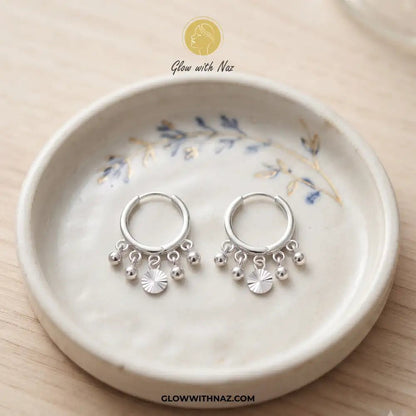 12mm Anti-Tarnish Silver 5 Drop Trending Hoops Earrings - 12mm Anti-Tarnish Silver 5 Drop Trending Hoops EarringsJewelryGlow With NazGlow With NazE045A pair of silver huggie hoop earrings with dangling charm beads and a small sunburst flower, displayed on a ceramic dish over a light wood background. Silver 3 Drop Separate Hoops, Glow With Naz earrings, stainless steel silver earrings, viral trending earrings, drop huggie hoops, silver dangle earrings, eye-catching drop hoops, waterproof silver j