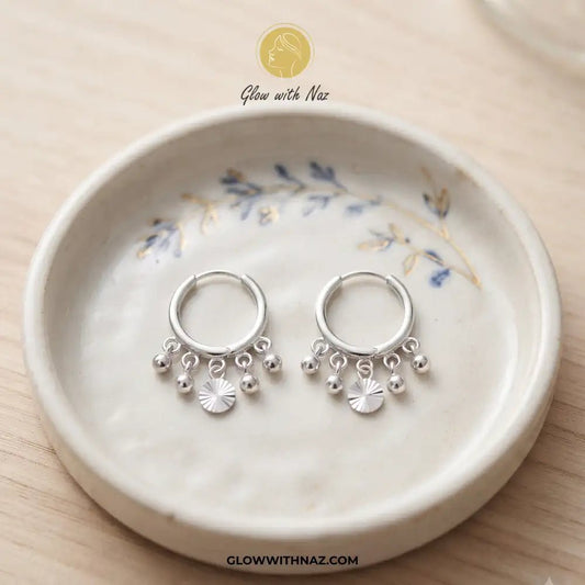 12mm Anti-Tarnish Silver 5 Drop Trending Hoops Earrings - 12mm Anti-Tarnish Silver 5 Drop Trending Hoops EarringsJewelryGlow With NazGlow With NazE045A pair of silver huggie hoop earrings with dangling charm beads and a small sunburst flower, displayed on a ceramic dish over a light wood background. Silver 3 Drop Separate Hoops, Glow With Naz earrings, stainless steel silver earrings, viral trending earrings, drop huggie hoops, silver dangle earrings, eye-catching drop hoops, waterproof silver j