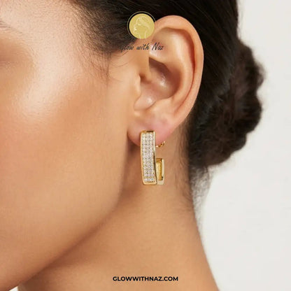 18K Gold Plated Diamond Square Geometric Statement Earrings - 18K Gold Plated Diamond Square Geometric Statement EarringsJewelryGlow With NazGlow With NazE053A close-up of a model's ear wearing a gold huggie hoop earring paved with small clear stones, with brand text 'Glow with Naz' and 'GLOWWITHAZ.COM' visible on the image.