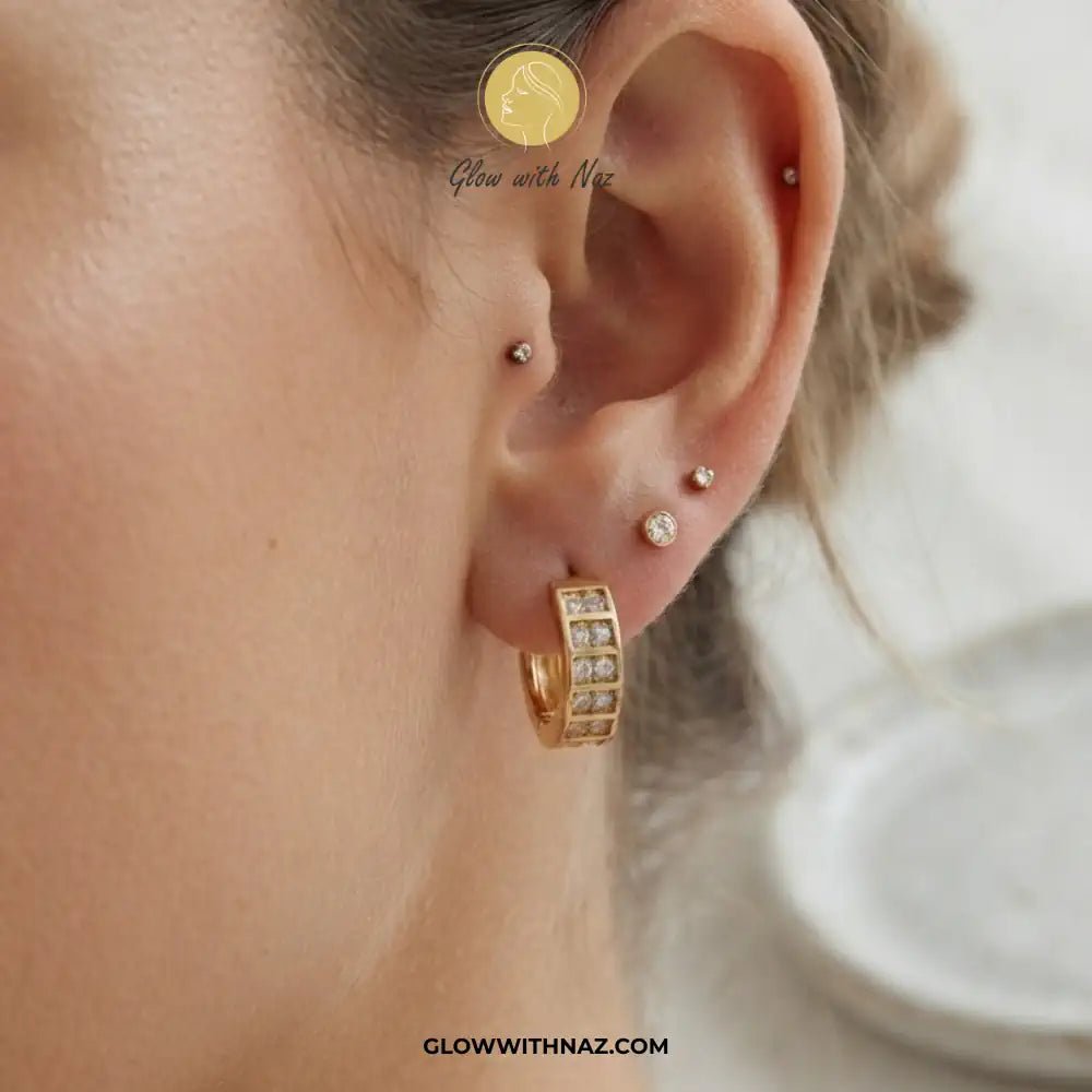 18K Gold Plated Sparkling Small 2-Line Diamond Huggie Earrings for Women - 18K Gold Plated Sparkling Small 2-Line Diamond Huggie Earrings for WomenJewelryGlow With NazGlow With NazE060A close-up of a model's ear showcasing a gold huggie hoop earring adorned with small clear crystals, alongside other ear piercings. The brand logo 'Glow with Naz' is visible as an overlay near the top.