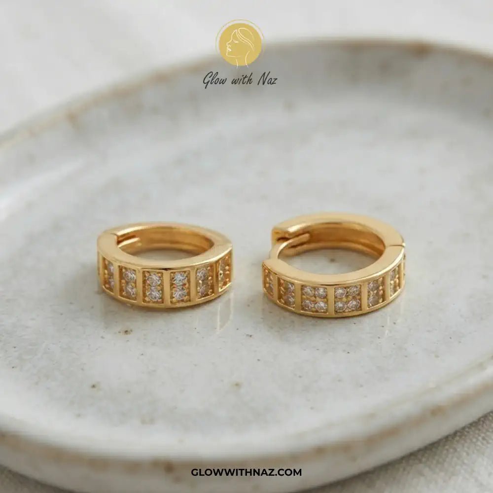 18K Gold Plated Sparkling Small 2-Line Diamond Huggie Earrings for Women - 18K Gold Plated Sparkling Small 2-Line Diamond Huggie Earrings for WomenJewelryGlow With NazGlow With NazE060A pair of gold huggie hoop earrings with rectangular pavé-set clear stones, displayed on a textured beige ceramic plate. The brand logo 'Glow with Naz' is visible at the top and the website 'GLOWWITHNAZ.