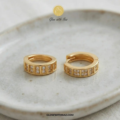 18K Gold Plated Sparkling Small 2-Line Diamond Huggie Earrings for Women - 18K Gold Plated Sparkling Small 2-Line Diamond Huggie Earrings for WomenJewelryGlow With NazGlow With NazE060A pair of gold huggie hoop earrings with rectangular pavé-set clear stones, displayed on a textured beige ceramic plate. The brand logo 'Glow with Naz' is visible at the top and the website 'GLOWWITHNAZ.