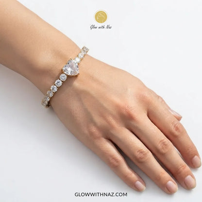A close-up of a person's hand and wrist wearing a delicate bracelet with a large pear-cut clear center stone and smaller clear stones along the band. The image has Glow with Naz branding on a clean white background.