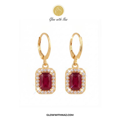 A pair of gold leverback drop earrings featuring rectangular emerald-cut red gemstones surrounded by small clear stones, displayed on a white background with the brand name Glow with Naz. 