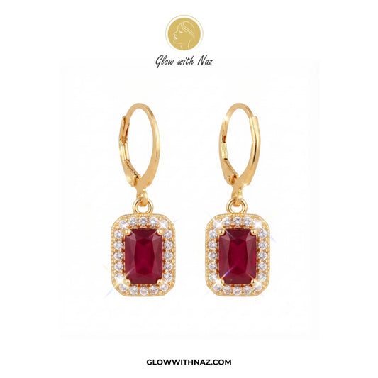 A pair of gold leverback drop earrings featuring rectangular emerald-cut red gemstones surrounded by small clear stones, displayed on a white background with the brand name Glow with Naz. 