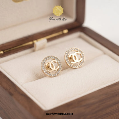 A pair of gold-toned stud earrings with a double-C logo design, surrounded by clear rhinestones, displayed in a cream-lined wooden jewelry box. A watermark logo for 'Glow with Naz' is visible at the top and bottom of the image. studs earrings, small earrings, girls earrings, simple earrings, earrings for girls, black earrings, earrings, ear rings, earrings tops, stud earrings, tops earrings, aesthetic earrings, hoop earrings, drop earrings, diamond earrings, pink earrings, green earrings, ear ring, earrings