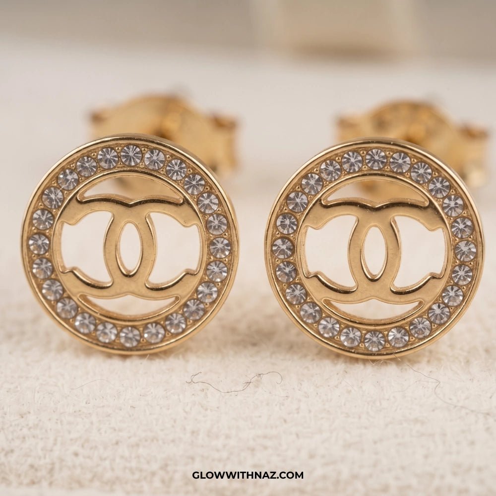 A pair of gold round stud earrings featuring a cutout interlocking double-C logo in the center, surrounded by a border of clear rhinestones, displayed on a textured cream surface. studs earrings, small earrings, girls earrings, simple earrings, earrings for girls, black earrings, earrings, ear rings, earrings tops, stud earrings, tops earrings, aesthetic earrings, hoop earrings, drop earrings, diamond earrings, pink earrings, green earrings, ear ring, earrings for women, flower earrings, golden earrings, ba