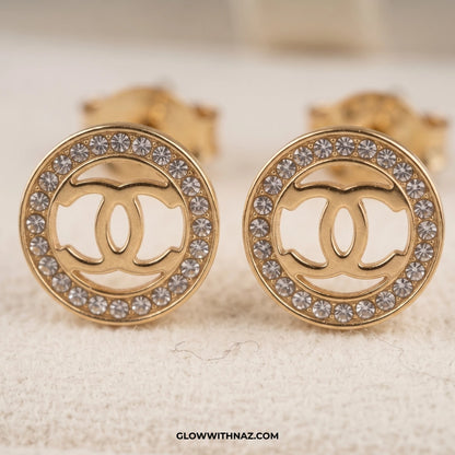 A pair of gold round stud earrings featuring a cutout interlocking double-C logo in the center, surrounded by a border of clear rhinestones, displayed on a textured cream surface. studs earrings, small earrings, girls earrings, simple earrings, earrings for girls, black earrings, earrings, ear rings, earrings tops, stud earrings, tops earrings, aesthetic earrings, hoop earrings, drop earrings, diamond earrings, pink earrings, green earrings, ear ring, earrings for women, flower earrings, golden earrings, ba