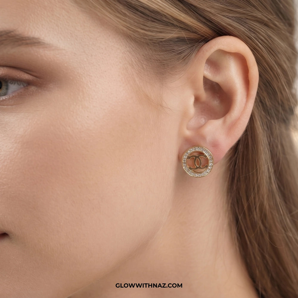 A close-up of a model's ear wearing a gold-toned stud earring with a double-C logo and a halo of small clear stones. The image is cropped tightly to the ear and side of the face, with a watermark at the bottom reading GLOWWITHNAZ. studs earrings, small earrings, girls earrings, simple earrings, earrings for girls, black earrings, earrings, ear rings, earrings tops, stud earrings, tops earrings, aesthetic earrings, hoop earrings, drop earrings, diamond earrings, pink earrings, green earrings, ear ring, earri