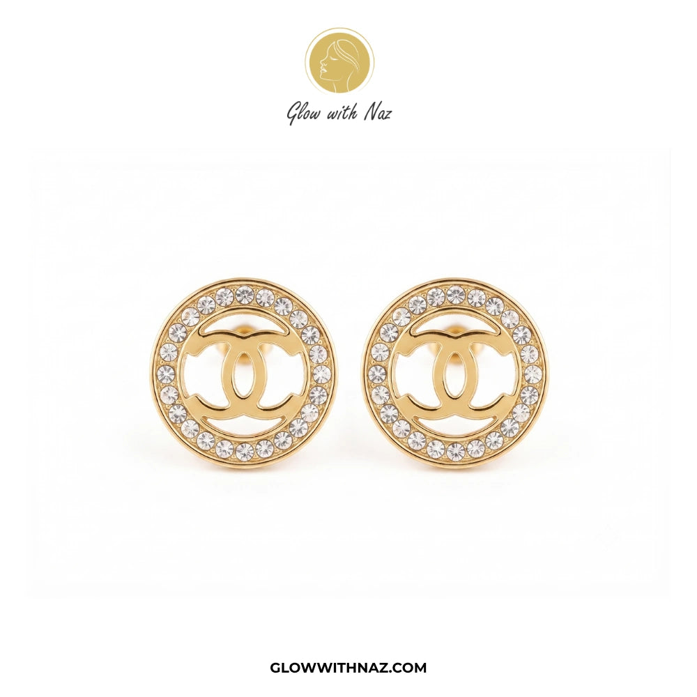 A pair of gold round stud earrings featuring a central interlocking CC logo surrounded by a border of clear crystals, displayed on a white background with a Glow with Naz logo at the top. studs earrings, small earrings, girls earrings, simple earrings, earrings for girls, black earrings, earrings, ear rings, earrings tops, stud earrings, tops earrings, aesthetic earrings, hoop earrings, drop earrings, diamond earrings, pink earrings, green earrings, ear ring, earrings for women, flower earrings, golden earr