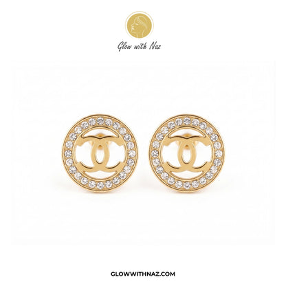 A pair of gold round stud earrings featuring a central interlocking CC logo surrounded by a border of clear crystals, displayed on a white background with a Glow with Naz logo at the top. studs earrings, small earrings, girls earrings, simple earrings, earrings for girls, black earrings, earrings, ear rings, earrings tops, stud earrings, tops earrings, aesthetic earrings, hoop earrings, drop earrings, diamond earrings, pink earrings, green earrings, ear ring, earrings for women, flower earrings, golden earr