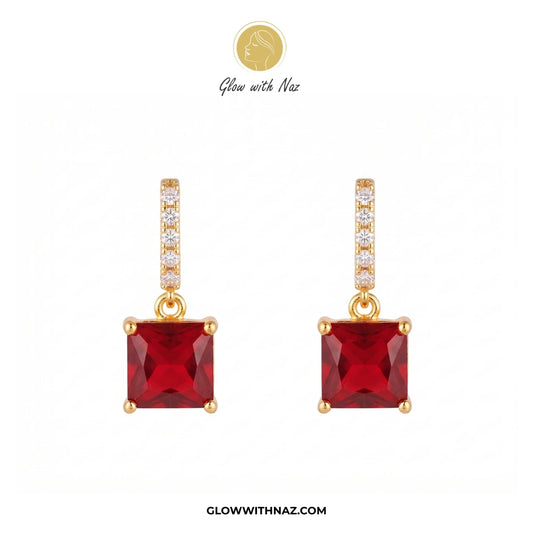 A pair of gold drop earrings featuring square-cut red gemstones, with a vertical bar of small clear stones above each red stone. The brand logo 'Glow with Naz' is visible at the top and the website 'GLOWWITHNAZ. studs earrings, small earrings, girls earrings, simple earrings, earrings for girls, black earrings, earrings, ear rings, earrings tops, stud earrings, tops earrings, aesthetic earrings, hoop earrings, drop earrings, diamond earrings, pink earrings, green earrings, ear ring, earrings for women, flow