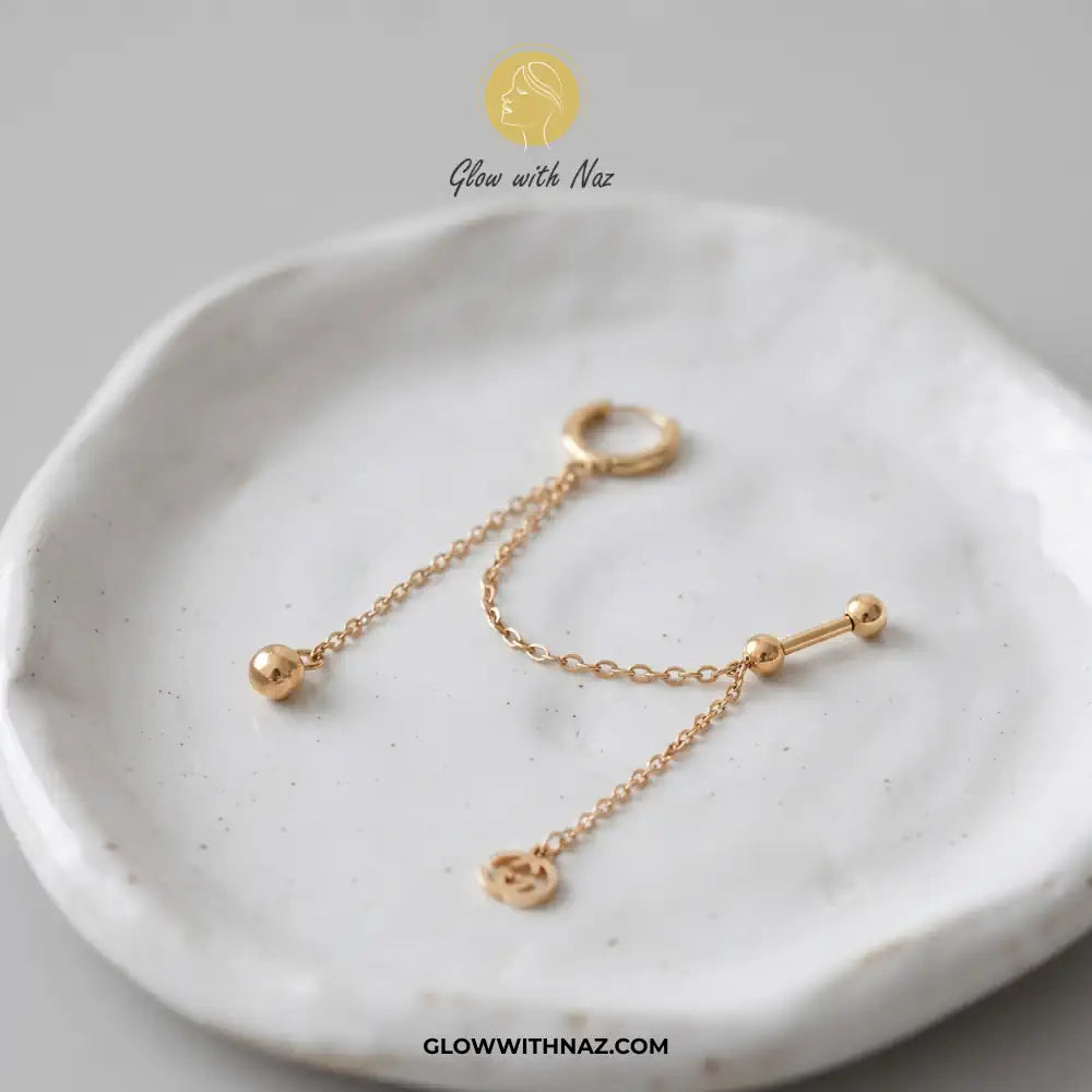 A pair of gold-toned ear piercings, including a hoop and a barbell with a dangling chain and small charm, displayed on a textured white ceramic dish. helix piercing, cartilage earring, double diamond stud, stainless steel jewelry, body jewelry, viral trending jewelry, ear stack earrings, cartilage piercing jewelry, minimalist piercing, tragus piercing, conch piercing, daith piercing, flat piercing, upper ear piercing, gold stainless steel piercing, sparkling helix stud, crystal cartilage jewelry, modern bod