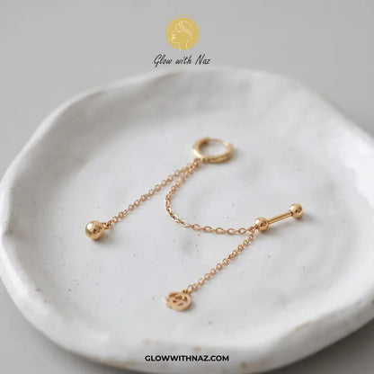 A pair of gold-toned ear piercings, including a hoop and a barbell with a dangling chain and small charm, displayed on a textured white ceramic dish. helix piercing, cartilage earring, double diamond stud, stainless steel jewelry, body jewelry, viral trending jewelry, ear stack earrings, cartilage piercing jewelry, minimalist piercing, tragus piercing, conch piercing, daith piercing, flat piercing, upper ear piercing, gold stainless steel piercing, sparkling helix stud, crystal cartilage jewelry, modern bod