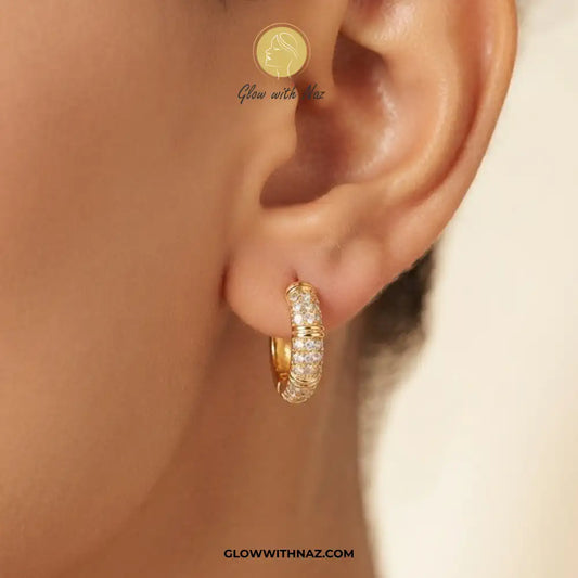 A close-up of a model's ear wearing a small gold huggie hoop earring paved with clear stones. The image features a watermark logo for 'Glow with Naz' and the website 'GLOWWITHNAZ.