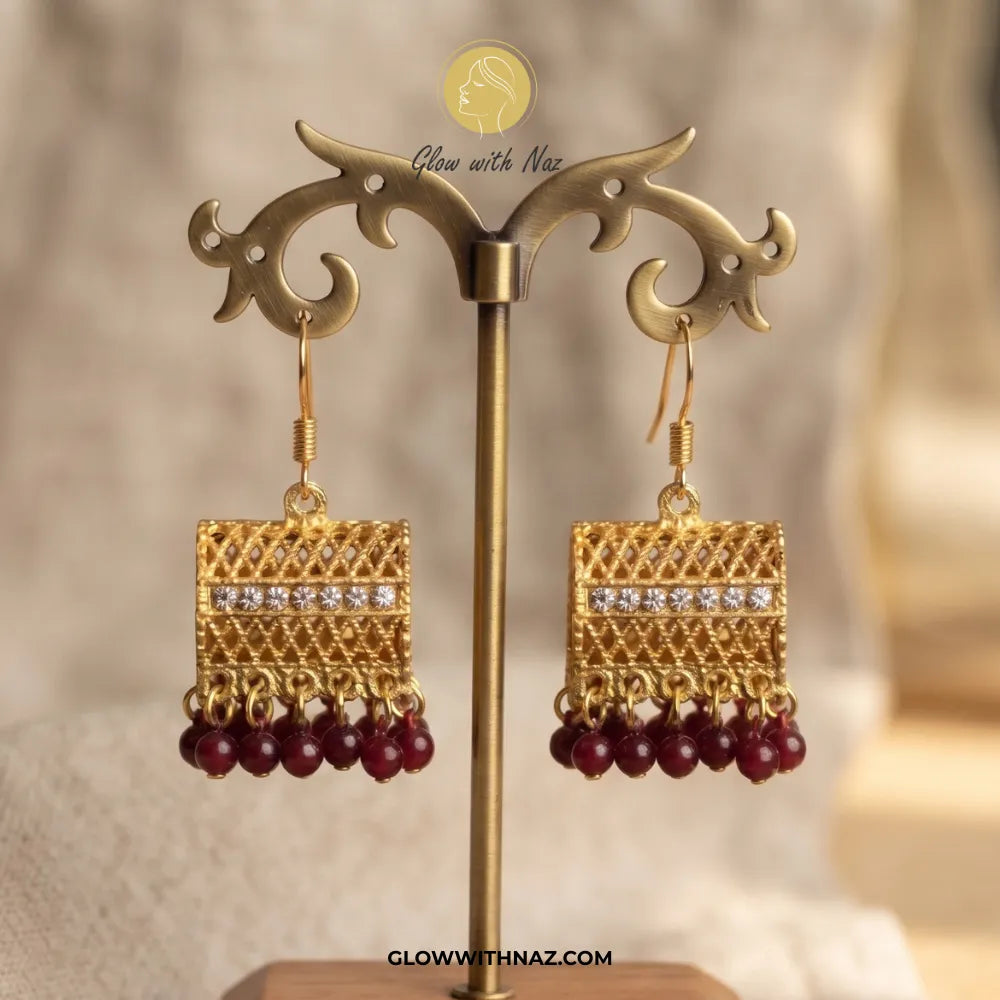 A pair of gold-toned traditional Indian jhumka earrings with red bead drops and a row of clear stones, displayed on a gold stand. The brand logo 'Glow with Naz' is visible at the top.