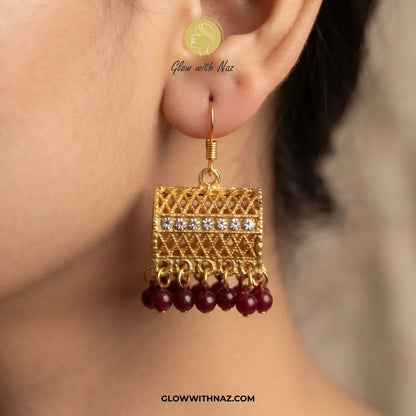 A close-up of a model's ear wearing a gold-toned traditional Indian jhumka earring with a rectangular filigree frame, a row of clear stones, and dangling maroon beads.