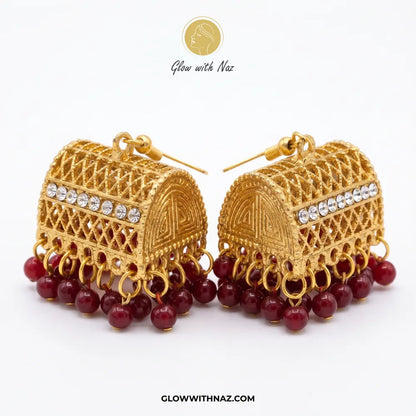A pair of traditional Indian gold jhumka earrings with red bead fringe and clear rhinestone accents, displayed on a clean white background with a brand logo at the top.