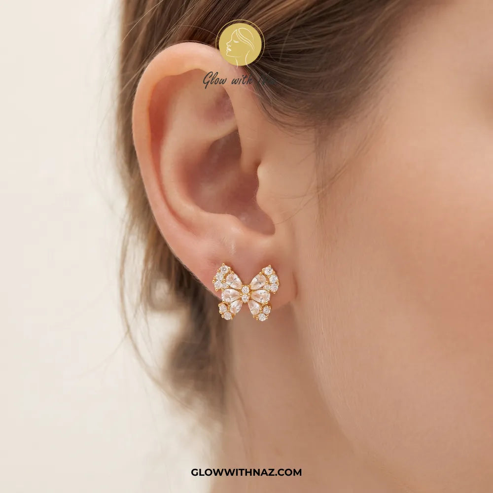 A close-up of a model's ear wearing a small bow-shaped stud earring with clear rhinestones on a gold-tone setting. The image includes a watermark logo for Glow with Naz.