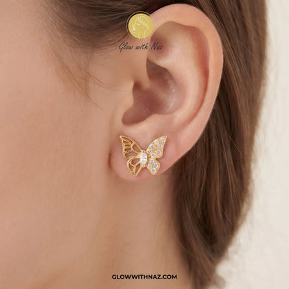 A close-up of a model's ear wearing a gold butterfly stud earring adorned with small clear crystals. The image features Glow with Naz branding.