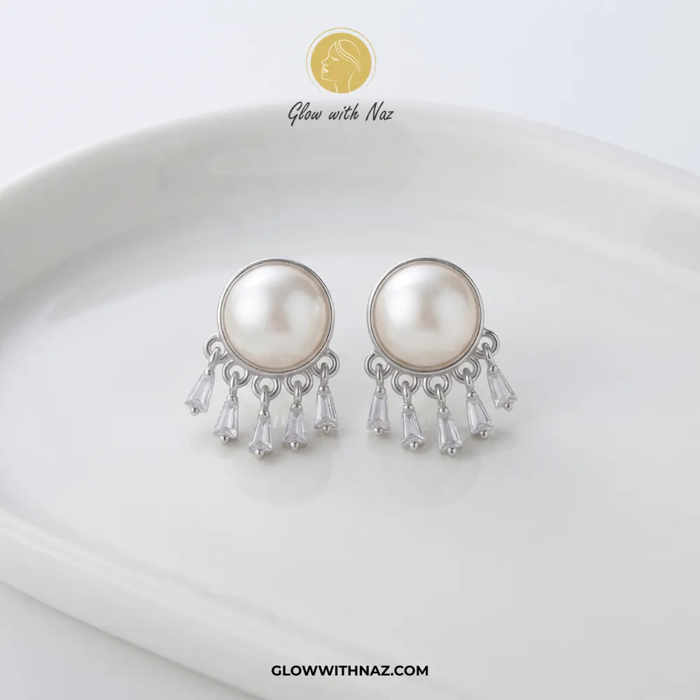 A pair of silver-toned dangle earrings with large round white faux pearls and clear teardrop crystal drops, displayed on a white ceramic dish. The brand logo 'Glow with Naz' is visible at the top and the website 'GLOWWITHNAZ.
