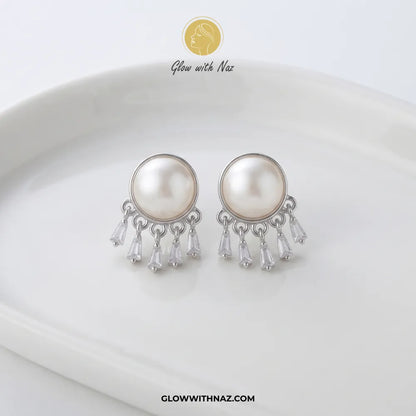 A pair of silver-toned dangle earrings with large round white faux pearls and clear teardrop crystal drops, displayed on a white ceramic dish. The brand logo 'Glow with Naz' is visible at the top and the website 'GLOWWITHNAZ.