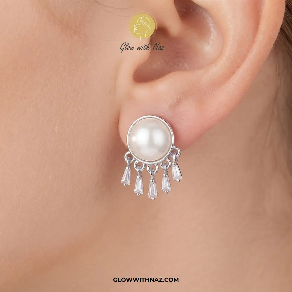 A close-up of a model's ear wearing a silver-toned dangle earring with a round white pearl and five dangling clear teardrop crystals. The brand logo 'Glow with Naz' is visible on the image.