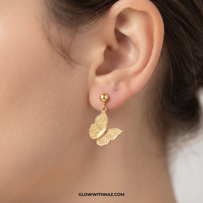 A close-up of a person's ear wearing a delicate gold butterfly drop earring with a small gold ball stud. The image features a watermark at the bottom reading 'GLOWWITHNAZ.
