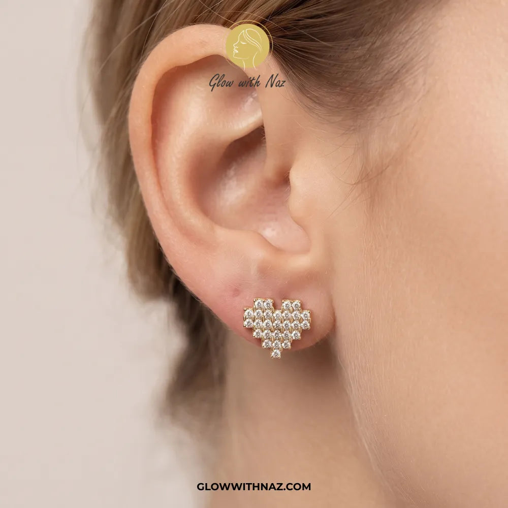 A close-up of a person's ear wearing a small heart-shaped stud earring made of clear rhinestones on a silver-toned base. The image has 'Glow with Naz' branding and a website watermark.