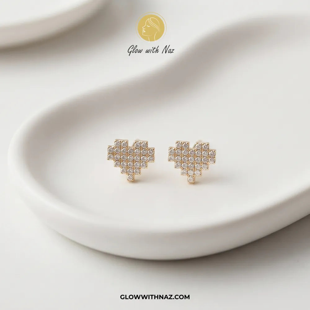 A pair of gold heart-shaped stud earrings covered in small clear crystals, displayed on a white ceramic dish. The brand logo 'Glow with Naz' is visible at the top, and the website 'GLOWWITHNAZ.