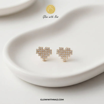 A pair of gold heart-shaped stud earrings covered in small clear crystals, displayed on a white ceramic dish. The brand logo 'Glow with Naz' is visible at the top, and the website 'GLOWWITHNAZ.