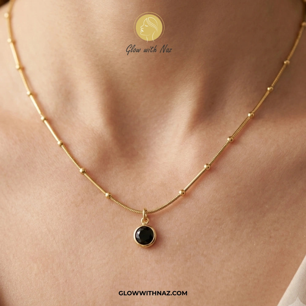 Gold necklace with a black pendant on a person's neck, branded 'Glow with Naz'. necklace, pendant necklace, necklace set, locket set, simple necklace, pendant set, pendant, chain necklace, diamond pendant, diamond necklace, locket necklace, neck chain, girls locket, heart locket, pendant for girls, necklace for girls, heart necklace, locket for girls, diamond locket, pendant necklace for girls, heart chain locket, stainless steel necklace, double chain necklace, ruby necklace, neck chain for girls, locket d