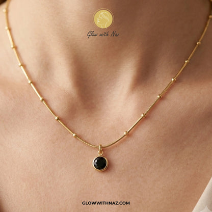 Gold necklace with a black pendant on a person's neck, branded 'Glow with Naz'. necklace, pendant necklace, necklace set, locket set, simple necklace, pendant set, pendant, chain necklace, diamond pendant, diamond necklace, locket necklace, neck chain, girls locket, heart locket, pendant for girls, necklace for girls, heart necklace, locket for girls, diamond locket, pendant necklace for girls, heart chain locket, stainless steel necklace, double chain necklace, ruby necklace, neck chain for girls, locket d