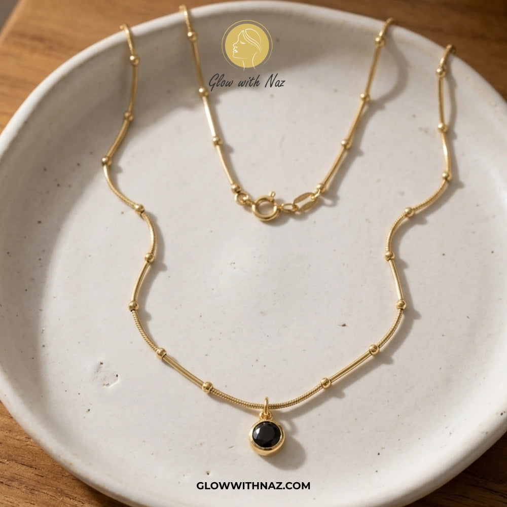 Gold necklace with a black pendant on a white ceramic dish, brand 'Glow with Naz' visible. necklace, pendant necklace, necklace set, locket set, simple necklace, pendant set, pendant, chain necklace, diamond pendant, diamond necklace, locket necklace, neck chain, girls locket, heart locket, pendant for girls, necklace for girls, heart necklace, locket for girls, diamond locket, pendant necklace for girls, heart chain locket, stainless steel necklace, double chain necklace, ruby necklace, neck chain for girl