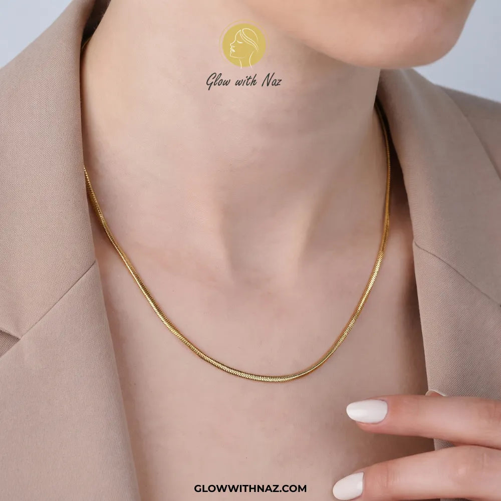 A close-up of a model's neck and upper chest wearing a delicate gold snake chain necklace. The model is wearing a beige blazer, and a watermark logo for 'Glow with Naz' is visible on the chest.