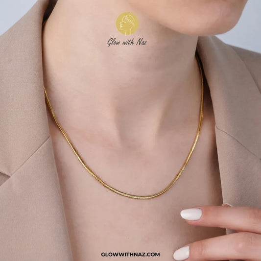 A close-up of a model's neck and upper chest wearing a delicate gold snake chain necklace. The model is wearing a beige blazer, and a watermark logo for 'Glow with Naz' is visible on the chest.