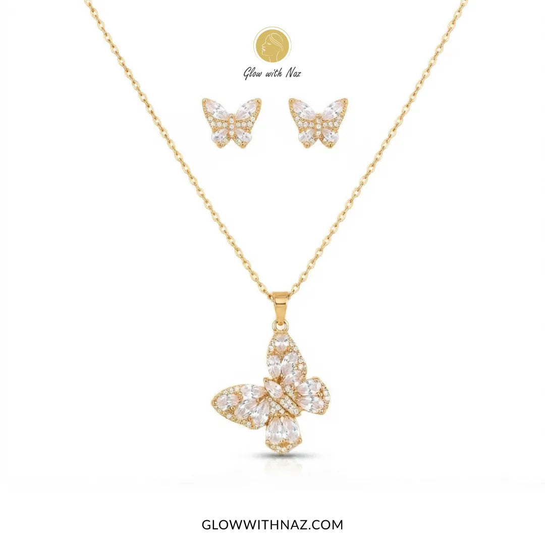 Gold butterfly pendant necklace and matching stud earrings set with clear crystals, displayed on a white background with a small inset showing the pendant glowing under Naz light.