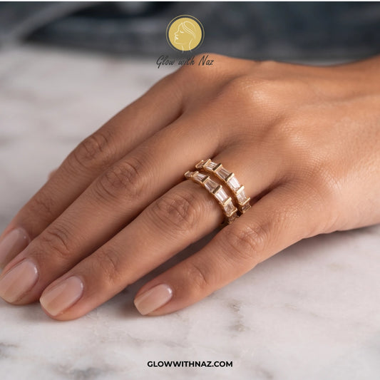 A close-up of a hand wearing two stacked gold rings with rectangular clear stones, resting on a light marble surface. A logo for Glow With Naz is visible at the top, and the website glowwithnaz. ring, girls rings, rings for girls, rings simple, simple rings, stainless steel jewelry, jewellery set, aesthetic jewelry, simple jewellery, chain, jewelry, artificial jewellery